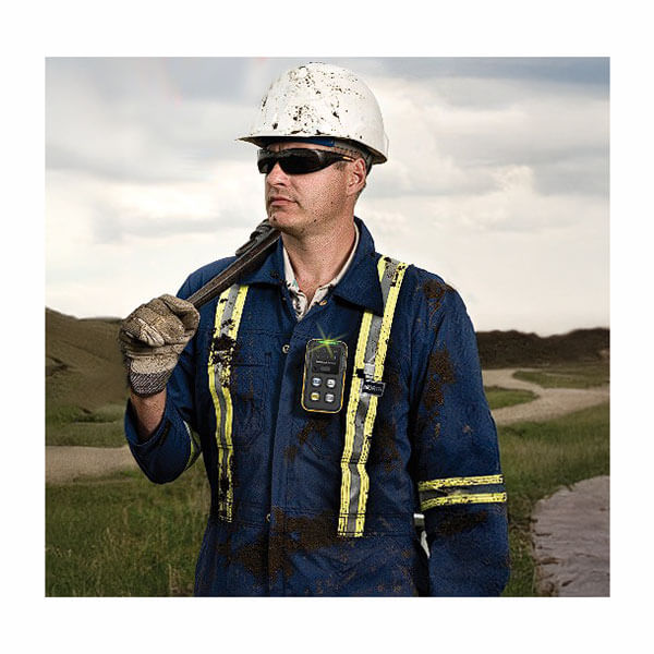 A construction worker in a blue jumpsuit with yellow stripes, white hard hat, gloves, and sunglasses stands outdoors holding a radio and the BW Technologies by Honeywell BW Icon+ Multi-Gas Detector. A dirt path and grassy field are behind him.