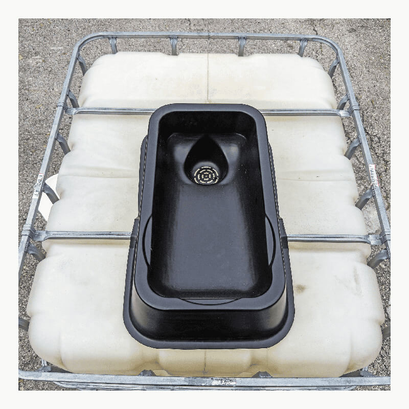 The UltraTech IBC Mini Funnel 0649, a black plastic sink with drain, sits atop a large white IBC with metal frame—creating a practical industrial liquid pouring system on a concrete surface.