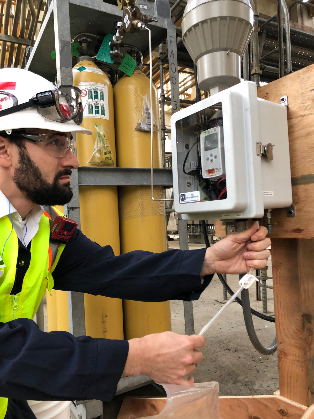 A male worker in safety gear adjusts an RKI Instruments GX-3R Pro 5 Gas Confined Space Monitor with Wireless Communication in an open control box at an industrial site, with gas cylinders and pipes visible in the background.
