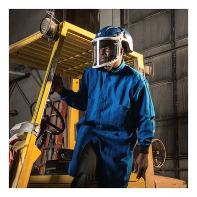 Dressed in a blue industrial coat and equipped with a Pureflo PureFlo 3000 PAPR Hard Hat, which includes a blue helmet and protective face shield, along with NIOSH-approved respirators for air-purifying safety, the individual stands next to a yellow forklift in an industrial setting.