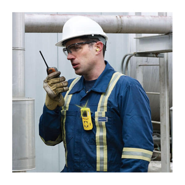 A worker in blue coveralls with PPE uses a walkie-talkie at an industrial site. A BW Honeywell GasAlert MicroClip XL Multi-Gas Monitor (MCXL-XWHM-Y-NA) by BW Technologies is clipped to his chest.