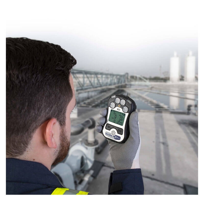 Wearing safety gear and gloves, a man uses a RAE Systems MicroRAE 4-Gas Diffusion Monitor M031-1111-200 to check readings at an industrial site with visible pipes and storage tanks in the background.