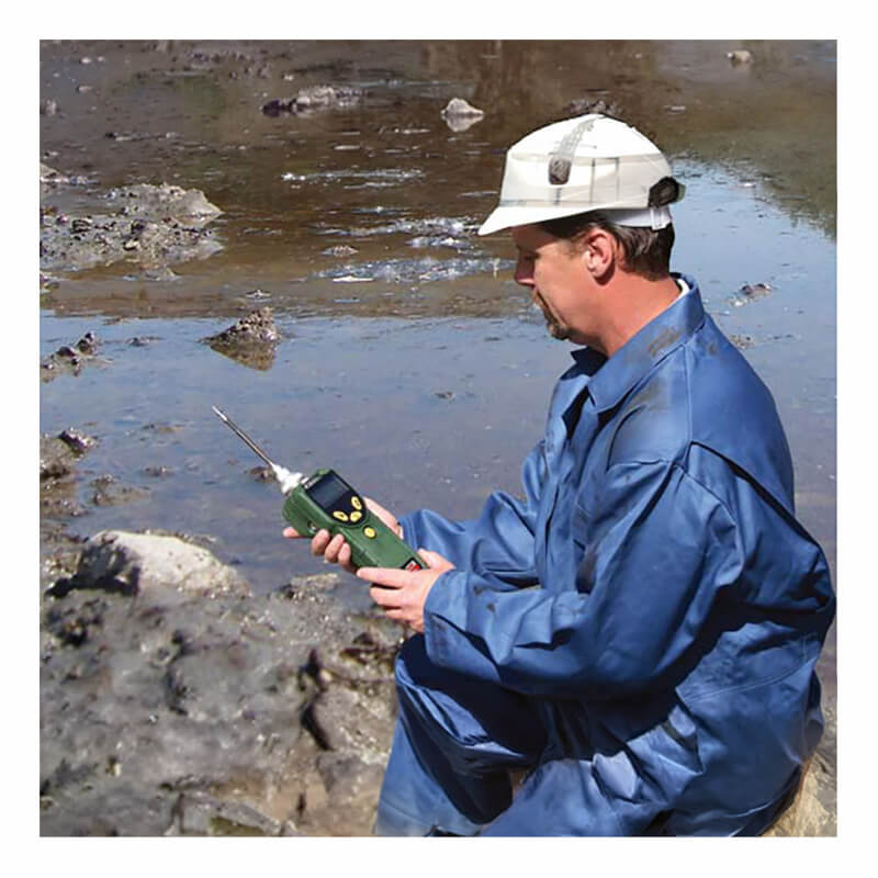 A man in blue coveralls and a white hard hat kneels by a muddy area, using the RAE Systems MiniRAE Lite PID Monitor 059-A to support environmental remediation efforts.