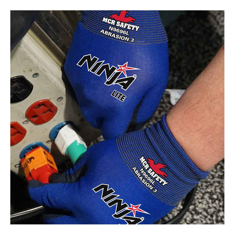 A person wearing blue MCR Safety Ninja Lite Work Gloves N9696, featuring a PU Coated Palm, is seen plugging a power cord into a socket. The gloves prominently display the "MCR Safety" and "Ninja Lite" logos. In the background, part of an electrical panel with red and white outlets can be seen, emphasizing the gloves' practicality for precision tasks.
