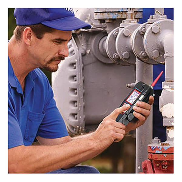A man in a blue shirt and cap uses the RKI Instruments GX-6100 4-Gas Monitor w/TC Sensor (72-61AXXT-C) to inspect industrial pipes and valves, checking the display near the machinery.