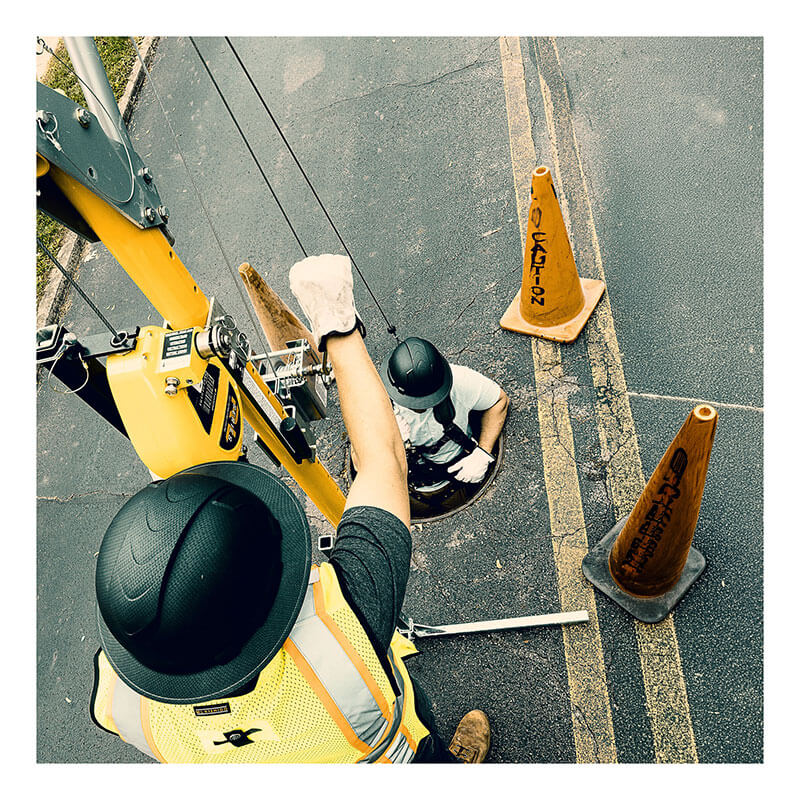 Two workers in safety gear, one on the street and one in a manhole using a French Creek 50ft Galvanized Rescue SRL R50G by French Creek Production, communicate amid orange cones. The scene is viewed from above.