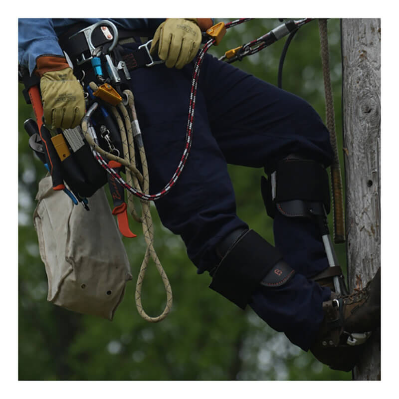 Wearing the NSA DRIFIRE Range Flex Pro Relaxed FR Work Pant in navy and arc-rated safety gear, a utility worker ascends a wooden pole with climbing equipment, tools hanging from their belt and ropes secured, set against a blurred green background.