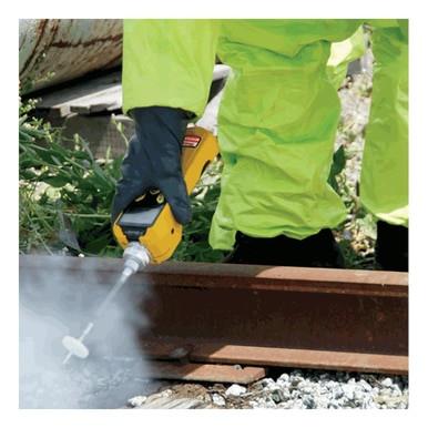 A person wearing a bright yellow protective suit and dark gloves is using a power tool, possibly with guidance from the RAE Systems MiniRAE 3000 PID, to apply a substance or perform maintenance on a rusty metal rail. Smoke or mist is visible around the work area, with small plants in the background.