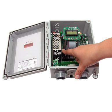 A hand is pointing at the circuit board inside an open gray electrical box, serving as a wall-mounted controller. Inside are various electronic components, including a digital display for gas detection. The cover is open, revealing a label that reads "RKI Beacon 110 Single Channel Wall Mount Fixed Systems Controller" by PK Safety Supply.