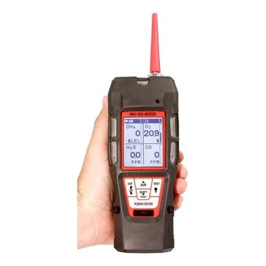 A hand holding an RKI Instruments GX-6000 PID Gas Monitor - Super Toxics 6 Gas Monitor, designed for confined space gas detection, displays various readings on its digital screen. This black and red device features a PID sensor, buttons at the bottom, and an antenna on top.