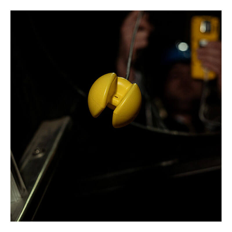 A close-up of the WatchGas Ball Float (Tubing NOT Included) SST-BALL-FLOAT by WatchGas, yellow and clam-shaped, is suspended by a wire with a blurred figure holding a yellow object in the dark background for confined space clearance.