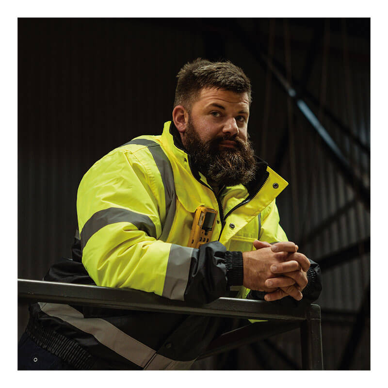 A bearded man wearing a high-visibility yellow and black safety jacket stands indoors by a metal railing in an industrial setting, showcasing the WatchGas SST4-MICRO Diffusion 4-Gas Detector for enhanced workplace safety.