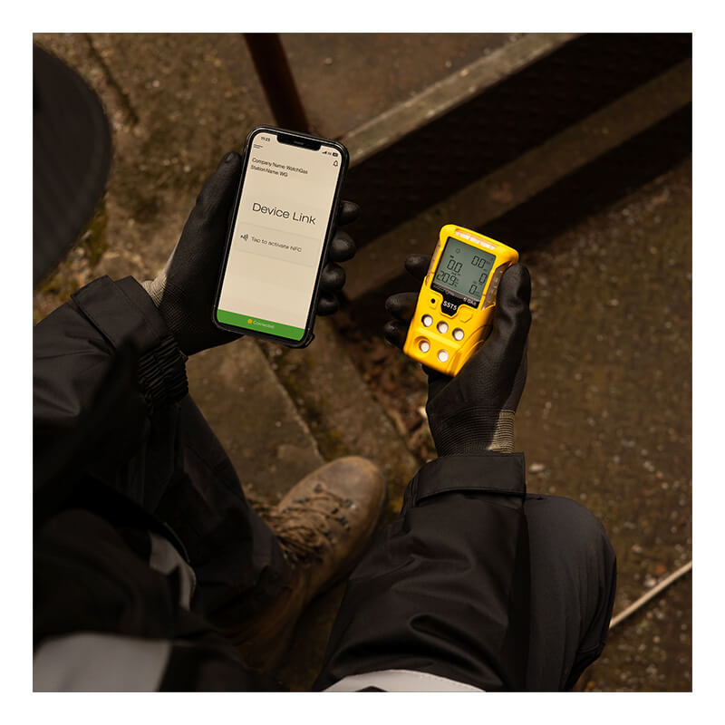 A person wearing gloves holds a smartphone displaying "Device Link" next to a yellow WatchGas SST5 Diffusion 5-Gas Detector (LPC/O2/CO/H2S/Exotic) by WatchGas while sitting outdoors on a metal staircase, with boots and industrial clothing visible.