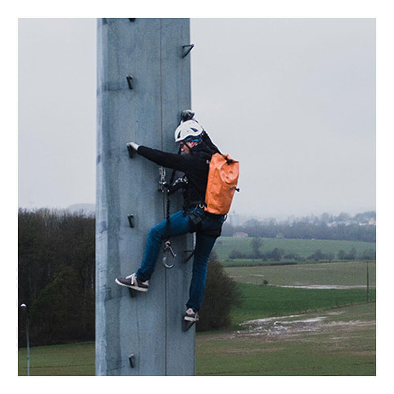A person using the Frontline Ladder Cable Vertical Lifeline System VLCS by Frontline Fall Protection climbs an outdoor vertical concrete structure with helmet, harness, and orange backpack; green fields and trees are visible in the background.