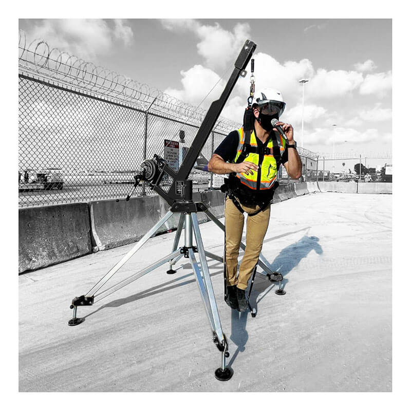 A worker in a safety helmet and high-visibility vest is suspended in a harness with Frontline Universal Suspension Trauma Straps ACTR by Frontline Fall Protection, attached to a tripod rescue system at a construction site with a fence and cloudy sky.