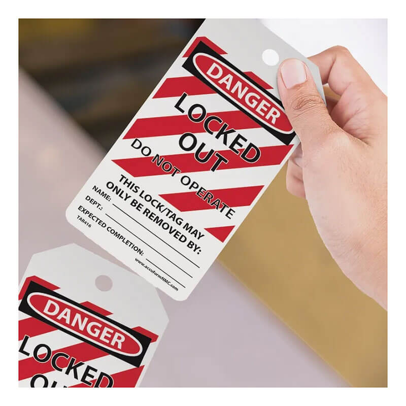 A hand holds an AccuformNMC OSHA Danger Tag By-The-Roll featuring bold red and black text that reads "Locked Out Do Not Operate." The tag, compliant with OSHA standards, includes sections for name, date, and expected completion on a background of diagonal red and white stripes.