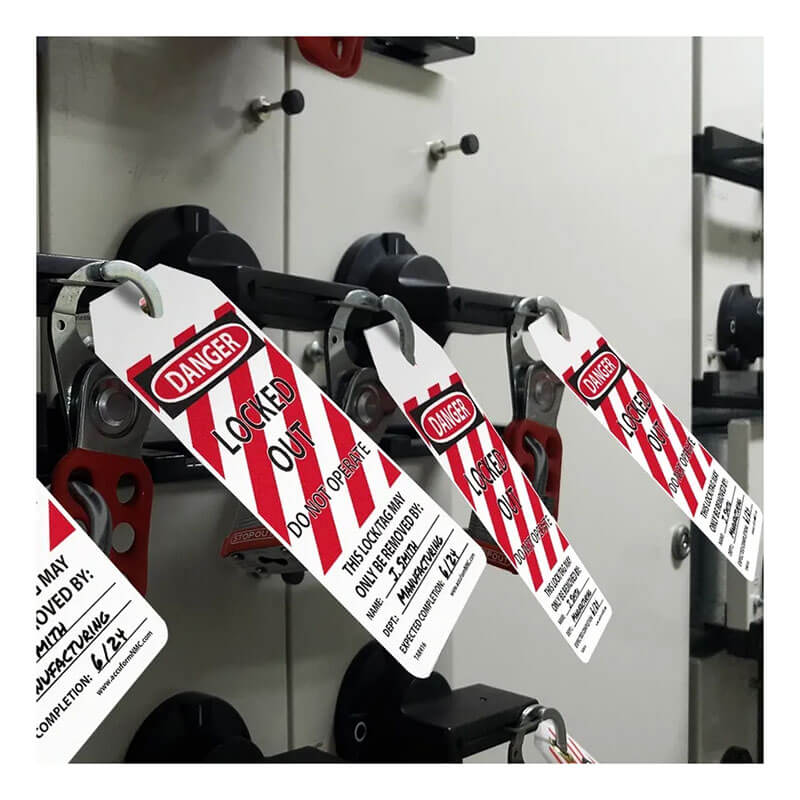 Close-up of several AccuformNMC OSHA Danger Tags By-The-Roll: Locked Out Do Not Operate (OSHA Style) attached to handles, featuring a "Danger: Locked Out, Do Not Operate" warning. The tags are designed with white and red stripes and have dedicated spaces for signatures and dates. The background displays a grey industrial panel, in compliance with OSHA safety standards.