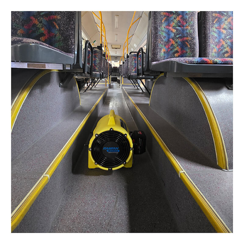 A Euramco RamFan UB20 18VDC blower, colored yellow, sits on the floor between rows of empty patterned seats inside a bus, facing toward the front.