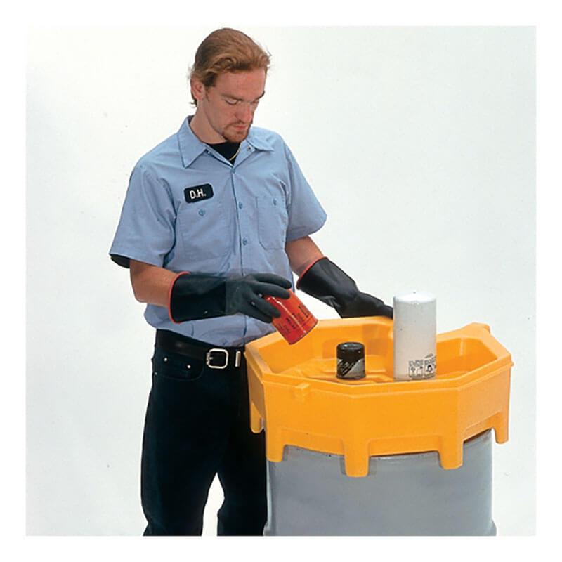 A person in a light blue shirt with a name tag, black gloves, and dark pants stands at a yellow spill containment drum. Using an UltraTech Global Funnel 049_, they pour liquid from a red container into the drum, ensuring precise liquid level monitoring with other small containers nearby.