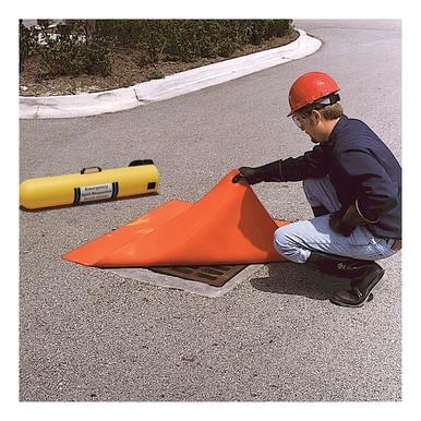 A worker in a hard hat and safety gear places an UltraTech Drain Seal from UltraTech International over a street drain, enhancing stormwater management. A yellow tool lies nearby on the paved road, framed by greenery in the background.