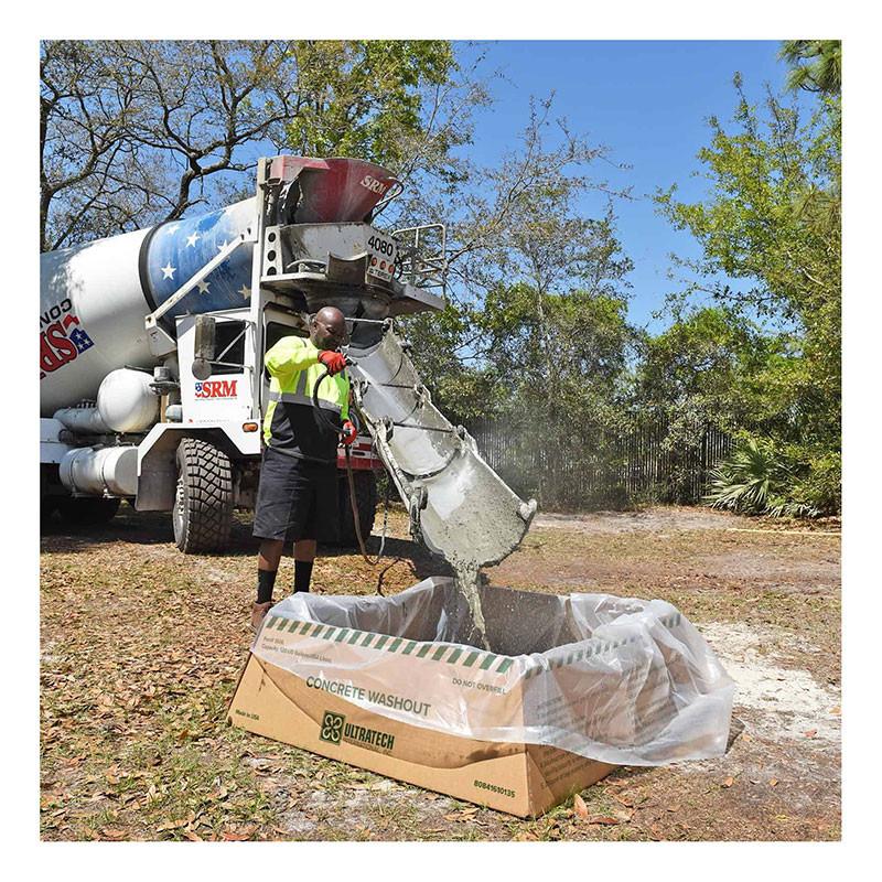 A worker in a neon vest and black shorts guides concrete from a cement truck chute into a lined cardboard container labeled "Concrete Washout," featuring the UltraTech Concrete Washout Berms - Economy Model 351. The setup complies with EPA regulations, and trees can be seen in the outdoor background.