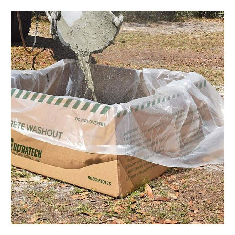 The UltraTech Concrete Washout Berms - Economy Model 351 is a cardboard box lined with clear plastic that serves as a portable concrete washout berm outdoors, ensuring EPA compliance. Labeled with UltraTech branding, it sits amid dry grass and soil as concrete is poured into it.