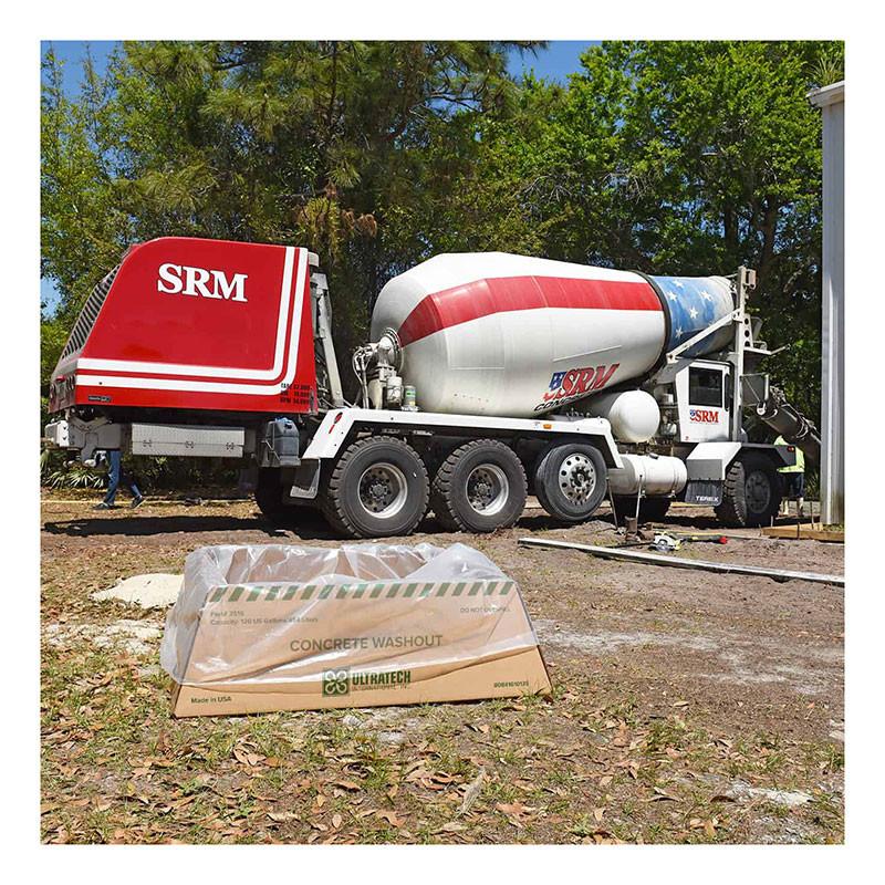 A vibrant red and blue concrete mixer truck with UltraTech's Economy Model 351 Concrete Washout Berm sits outdoors. The cardboard box indicates EPA compliance, surrounded by trees and grass under the clear sky, ensuring eco-friendly practices at work.