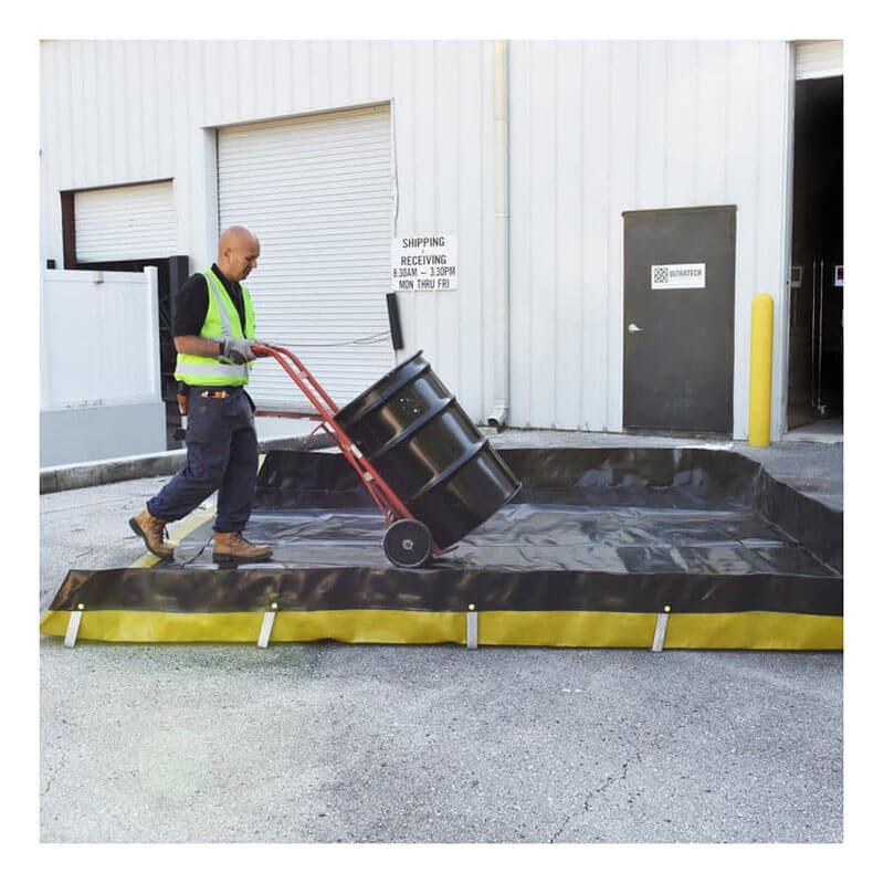 Wearing a safety vest, a man maneuvers a large black barrel with a red hand truck outside the warehouse. He places it into an UltraTech Ultra-Containment Berm Economy PE Model 824_ on the concrete by the roll-up doors, prioritizing spill containment.