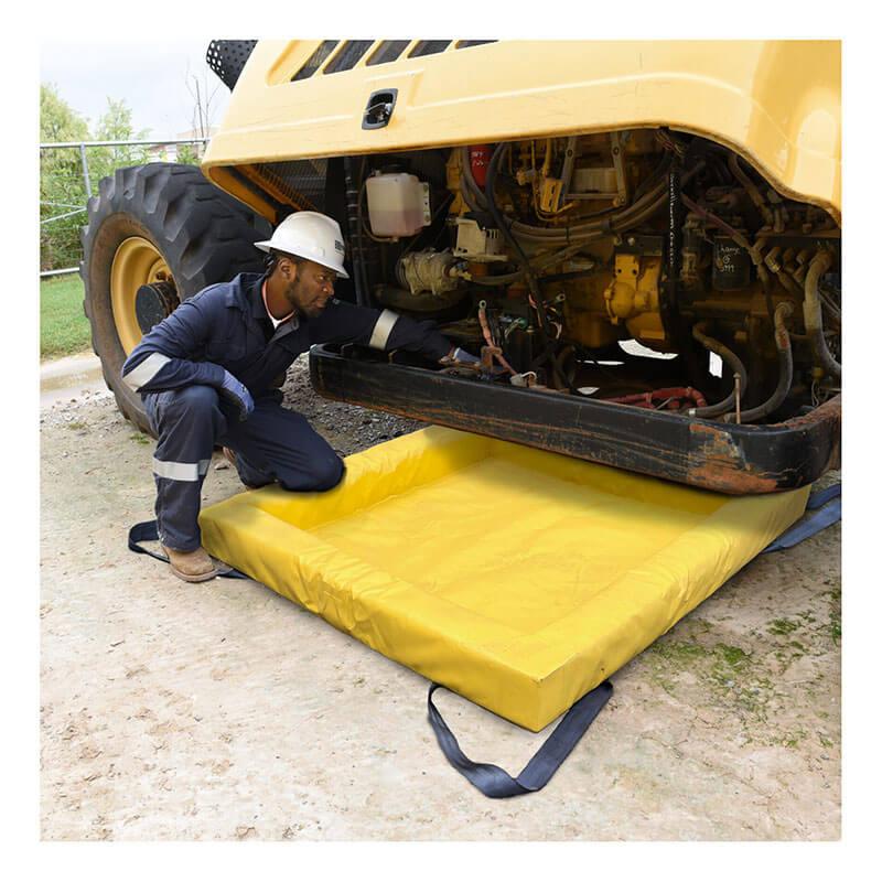 A worker in safety gear kneels on a yellow UltraTech Containment Berm - Foam Wall XC Model 898 spill containment mat while inspecting the underside of a large yellow construction vehicle.
