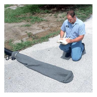 A person in a blue uniform kneels on a paved surface near grass, jotting notes in a notebook. In front is an UltraTech Oil & Sediment Model Pipe Socks 970_-OS, a gray covered object with a black hose for sediment and oil control.