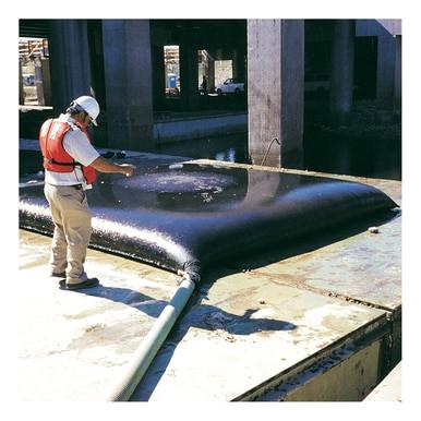 A worker in a hard hat and orange vest inspects an UltraTech Dewatering Bag 972_-OS, inflated and connected to a green hose. The industrial scene, featuring concrete structures and water, indicates storm water pollution prevention efforts are in progress.