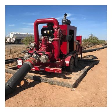 A large red industrial pump on a trailer is connected to black hoses in a dirt area outdoors, with UltraTech Foam Wall Model Containment Berms by UltraTech set up nearby. Trees and a truck are in the background under a clear blue sky.
