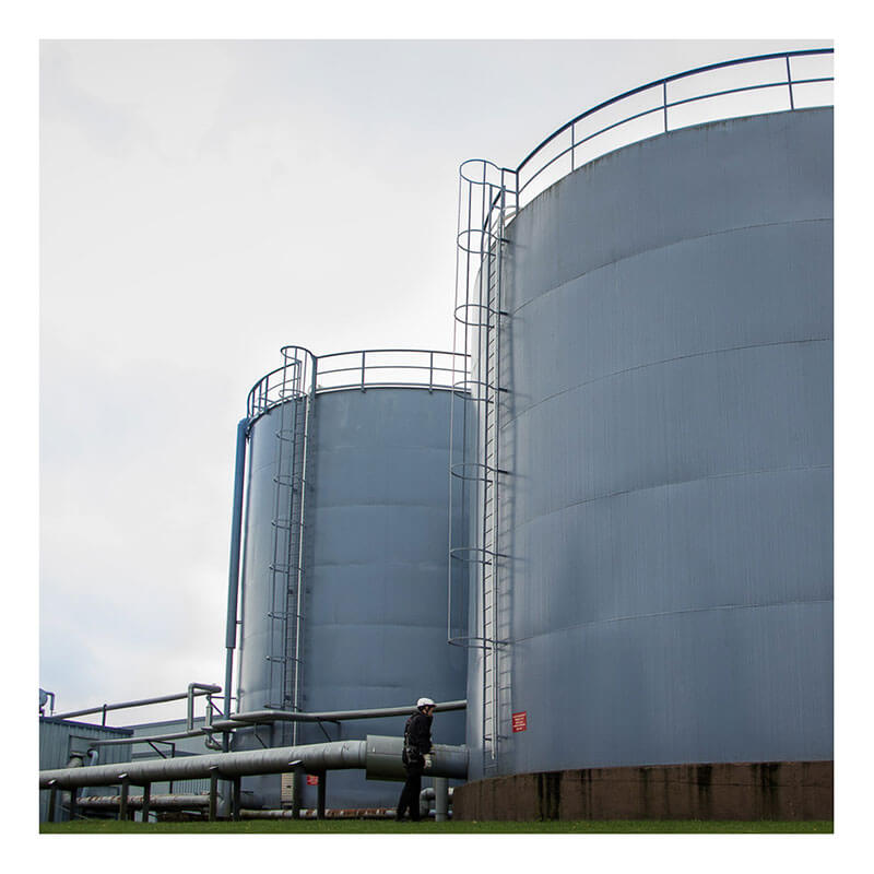 Two large cylindrical metal storage tanks with roof access ladders and railings equipped with the Frontline Securail Pro Vertical Lifeline System VLRS by Frontline Fall Protection stand outdoors as a person in hard hat and safety gear walks nearby.