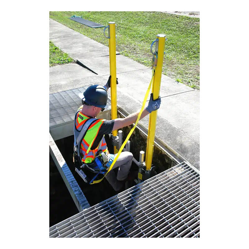 A worker in safety gear is tethered while descending into a confined space using the French Creek Apex Retractable Ladder Anchor Post by French Creek Production, next to a metal grate and grass-lined sidewalk.
