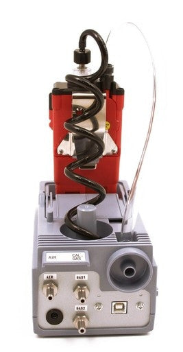 A close-up of a gray and red electronic device, resembling the RKI SDM-2012 Calibration Station for GX-2012 from PK Safety Supply, with various ports and connectors. A coiled black cable extends upwards, flanked by labels near the input ports. The device features a metallic and plastic exterior for rugged durability.