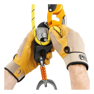 A person wearing tan and brown gloves is demonstrating climbing equipment, highlighting a yellow and black rope designed for rope access. The setup includes an orange carabiner and a Petzl RIG Compact Self-Braking Descender (D021AA00). The focus is entirely on the hands and equipment, with no background visible.