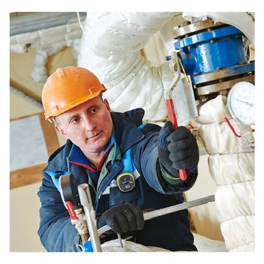A worker in an orange hard hat and blue jacket uses a wrench to adjust equipment, surrounded by insulated pipes and a pressure gauge, while the Honeywell BW Solo Wireless Gas Detector H2S from BW Technologies ensures safety.