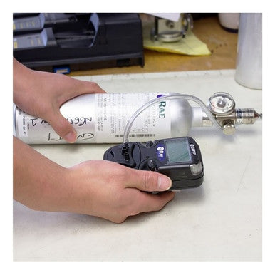 An individual is holding a PK Safety Supply's RAE Systems Gas Monitor Calibration Service, connected to a calibration gas cylinder. The display screen on the monitor and the valve and pressure gauge on the cylinder are clearly visible. They are situated on a tabletop amidst various equipment, guaranteeing accurate calibration of the gas detector.