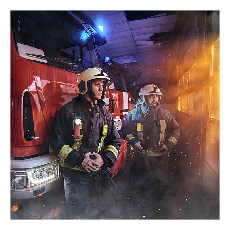 Two firefighters in full gear stand indoors by a fire truck, smoke and embers swirling, as their BW Technologies by Honeywell BW Clip 2 Year O2 Single Gas Detector BWC2-X ensures air safety amid orange and blue emergency lights.