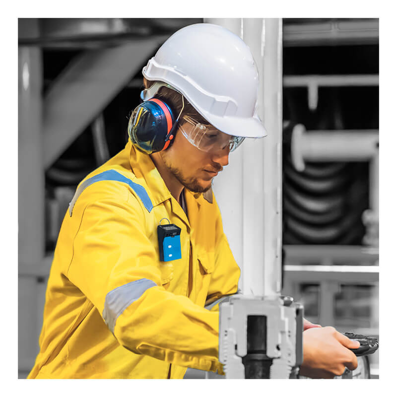 A worker equipped with a white helmet, safety glasses, ear protection, and a yellow protective suit operates industrial equipment indoors while using the GfG G222E Micro 5 H2S Single Gas Detector (model G222E-0200) from GfG.
