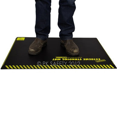 A person in brown work boots and blue jeans stands on a Working Concepts ErgoKneel Extreme Standing Mat 5032, featuring a black surface with yellow caution stripes. This anti-fatigue mat, made from closed cell foam pad material, provides comfort for long periods of standing while working.