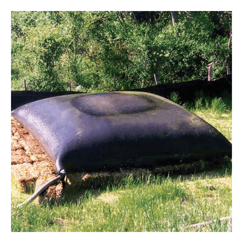 An UltraTech Dewatering Bag 972_-OS (black, large) sits on grass with a hose attached, surrounded by hay bales. Lush green trees and shrubs in the background show effective stormwater pollution prevention by UltraTech.
