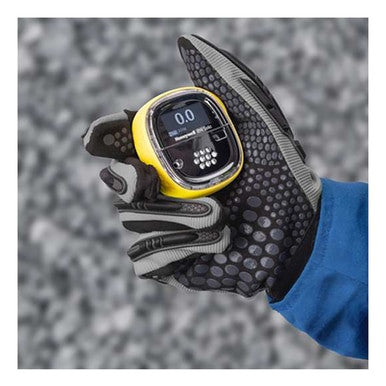 A gloved hand holds a Honeywell BW Solo Wireless Gas Detector H2S ext range BWS1-H2-Y displaying "0.0." The glove is patterned in black and gray, with a blue sleeve visible, emphasizing safety. A blurred gray background enhances the scene's subtle intensity. This detector is by BW Technologies by Honeywell.