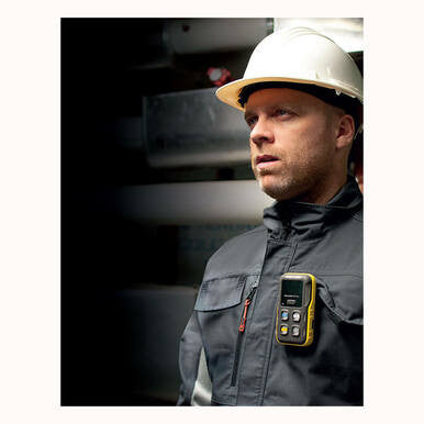 A man in a hard hat and work jacket stands indoors, intently holding a BW Honeywell 4-Gas Detector (LEL, O2, H2S, CO) ICON-Y-H attached to his chest. The sleek device softly glows amidst the dimly lit industrial backdrop as he looks to the side with focus.