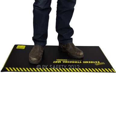 A person stands on a WORKING CONCEPTS ErgoKneel Extreme Standing Mat 5030, which features "Extreme Standing Mat" in yellow letters. This standing mat, known for its yellow-and-black striped edges, is placed against a white background. The person wearing jeans and brown work boots appreciates the comfort provided by the nitrile foam cushioning.