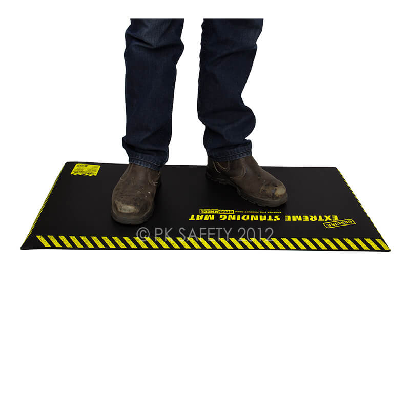 A person stands on a WORKING CONCEPTS ErgoKneel Extreme Standing Mat 5030, which features "Extreme Standing Mat" in yellow letters. This standing mat, known for its yellow-and-black striped edges, is placed against a white background. The person wearing jeans and brown work boots appreciates the comfort provided by the nitrile foam cushioning.