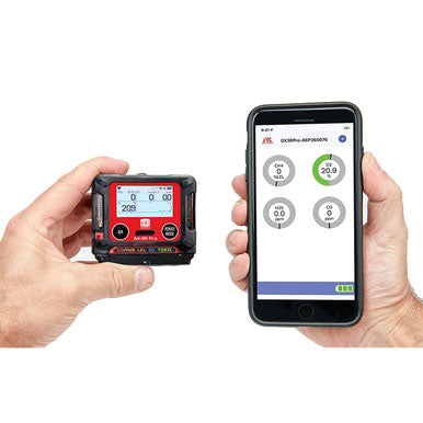 Two hands hold devices: the left grips an RKI GX-3R Pro Gas Confined Space Monitor with a red digital display, while the right holds a smartphone displaying the connected app via Bluetooth communication, featuring circular gauges and numerical readings.