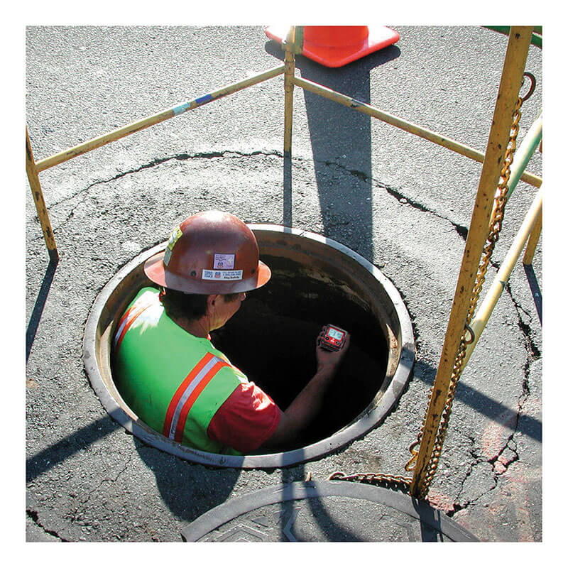 A worker in a hard hat and safety vest descends into a manhole, holding the RKI Instruments GX-3R 4 Gas Confined Space Monitor (72-RA-C). Safety barriers and an orange cone surround the area on the pavement.