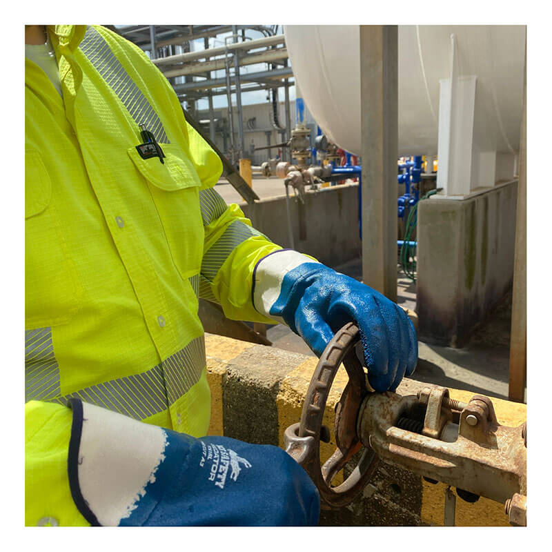 A person in a bright yellow safety jacket and MCR Safety Memphis Predator Rough Fully Coated Nitrile Cuff Gloves 9761R is turning a metal valve at an industrial site with visible pipes and large tanks in the background.