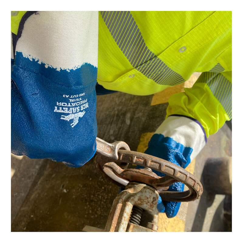 Wearing MCR Safety Memphis Predator Rough Fully Coated Nitrile Cuff Gloves 9761R, a person in a high-visibility yellow jacket grips and turns a rusted metal valve wheel.